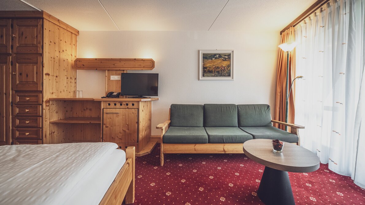 Double room with red carpet, window and curtains, sofa with small table, pine wood interior | © Davos Klosters Mountains 